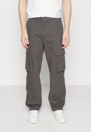 Persona con pantalones cargo gris oscuro con bolsillos de solapa y zapatillas blancas, de pie frente a un fondo claro y liso.