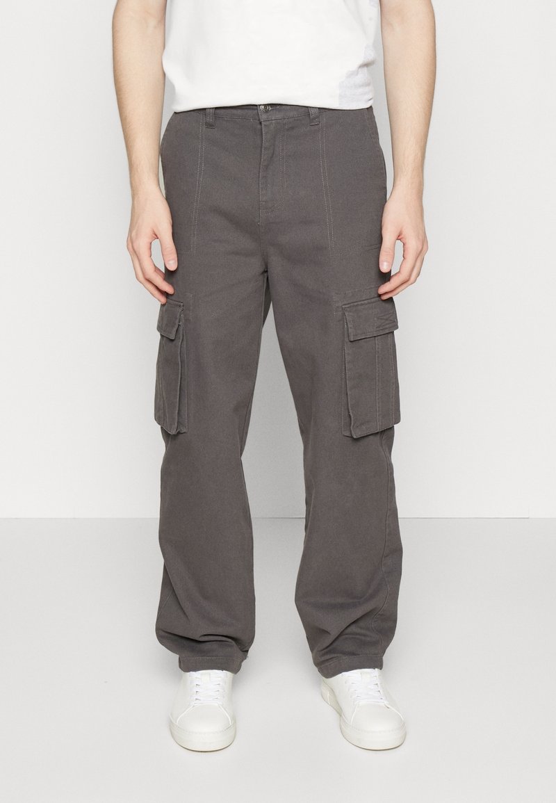 Personne portant un pantalon cargo gris foncé avec des poches à rabat et des baskets blanches, debout devant un fond clair uni.