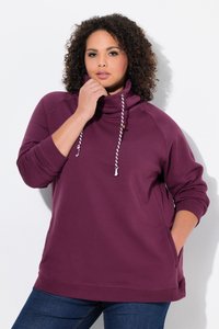 Femme aux cheveux bouclés portant un pull violet à col montant avec des cordons blancs, une main touche le col, l'autre dans la poche.