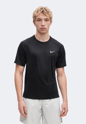 Mladý muž čelí dopredu, oblečený v čierne tričko Nike s krátkym rukávom a svetlé krátke nohavice na jednoduchom pozadí.