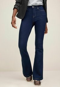 Jeans in denim a gambale svasato di colore blu scuro con dettagli di cucitura visibili. Il tessuto ha una texture liscia, completata da una classica chiusura con bottone.