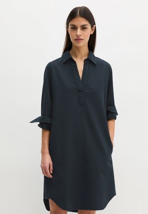 Femme portant une robe chemise ample bleu foncé à manches longues retroussées, debout devant un fond clair uni.