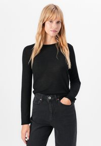 Ξανθιά γυναίκα φορώντας μαύρη μπλούζα με μακριά μανίκια και σκούρα ψηλόμεσα τζιν, στέκεται με το ένα χέρι στην τσέπη μπροστά σε λευκό φόντο.