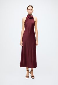 Robe bordeaux à encolure américaine avec un col bénitier, texture lisse et silhouette longue jusqu'au sol, associée à des escarpins noirs à talons aiguilles.