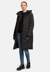 Cappotto lungo imbottito nero con cappuccio, chiusura a zip e bottoni automatici, caratterizzato da un motivo trapuntato e tasche laterali, indossato sopra un maglione nero e jeans.
