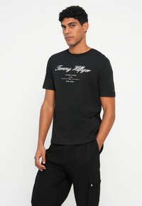 Tricou negru din bumbac cu text alb "Tommy Hilfiger", mâneci scurte și design cu guler rotund, asortat cu pantaloni cargo negri.