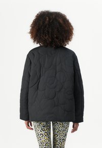 Giacca nera trapuntata con texture floreale, scollatura rotonda e maniche lunghe, abbinata a leggings a fantasia nelle tonalità di giallo e grigio.