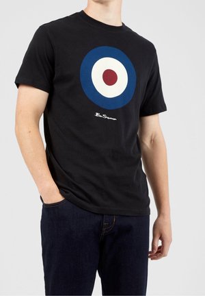 Mężczyzna ubrany w czarną koszulkę z niebiesko-biało-czerwonym okrągłym wzorem celownika oraz napisem "Ben Sherman", połączoną z ciemnoniebieskimi dżinsami.