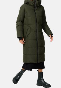 Olivengrøn puffermantel med hætte, udstyret med lynlåslommer, en lynlåslukning foran og et quiltet mønster, kombineret med sorte ankelstøvler.