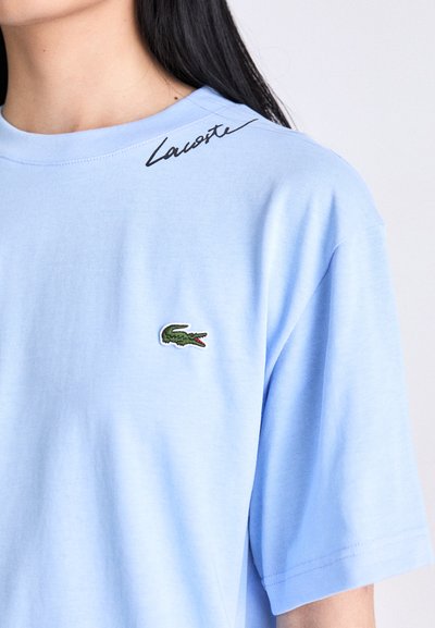 Жена, облечена с светлосиня тениска на Lacoste с зелен логотип на крокодил на гърдите и черен надпис на Lacoste в близост до деколтето.