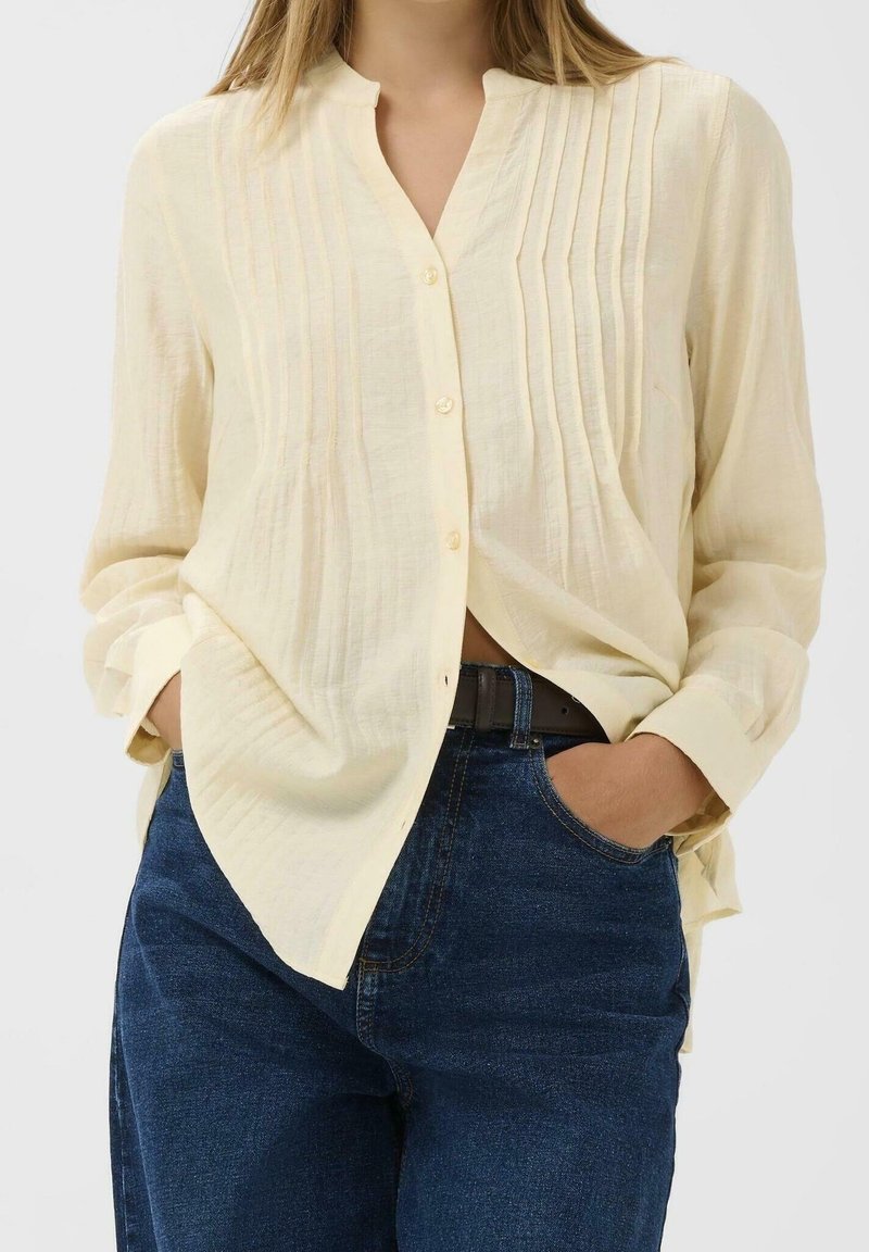 Femme portant un chemisier ample jaune clair à boutons avec des plis verticaux, légèrement rentré dans un jean bleu foncé, les mains dans les poches.