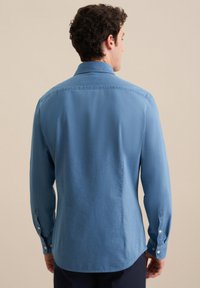 Uomo con capelli ricci corti che indossa una camicia a maniche lunghe azzurro chiaro aderente, in piedi di spalle contro uno sfondo beige uniforme.