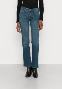 Jeans de denim azul con cintura ajustada, piernas acampanadas y un acabado ligeramente deslavado. Cuenta con cinco bolsillos y un cierre de botón de marca.