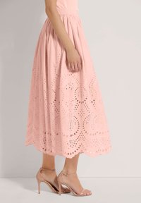 Rosa Midikleid mit gestanzter Stickerei, das kreisförmige Muster und einen gewellten Saum aufweist. Kombiniert mit nackten High-Heels.