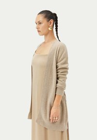 Beige stickad kofta med texturerat mönster, öppen front och långa ärmar; bärs över en enkel beige klänning. Har en jämn yta.