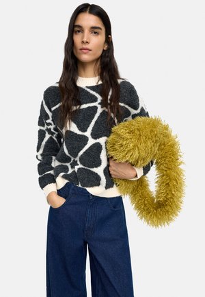 Kobieta z długimi ciemnymi włosami nosi czarno-biały sweter w wzory oraz szerokie jeansy, trzymając dużą puszystą żółtą szalik.