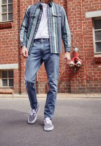 Randig grön och vit skjorta med knappar över en vit t-shirt, tillsammans med blå jeans och grå sneakers, mot en tegelbakgrund.