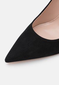 Chaussure à talon aiguille en daim noir avec un bout pointu et un design en découpe. Présente un intérieur en cuir lisse et clair ainsi que des détails de couture subtils.