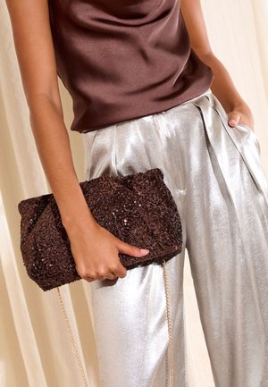 Smeđa večernja torbica s lancem, ukrašena sjajnim šljokicama i uvijenim dizajnom. Model nosi srebrne, sjajne hlače širokih nogavica s naborima i smeđu majicu.