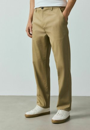 Mand iført beige bukser med lige pasform og hvide sneakers står på en grå gulv mod en ensfarvet lys baggrund.