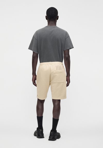 Homme debout de dos, portant un t-shirt gris foncé délavé, un short beige avec une poche arrière, des chaussettes noires et des chaussures de sport noires.