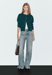 Jonge vrouw in een donkergroene knooptrui, wit topje, wijdvallende vervaagde jeans, zwarte sandalen en een donkerbruine handtas in haar hand.