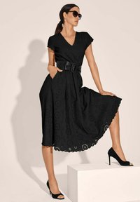 Schwarzes V-Ausschnitt-Kleid mit kurzen Ärmeln, Lochmuster, gewelltem Saum, tailliert mit Gürtel, kombiniert mit schwarzen High-Heels und Sonnenbrille.