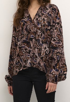 Femme portant un chemisier à manches longues à motif cachemire foncé avec manches bouffantes et pantalon noir ajusté, debout devant un fond uni.