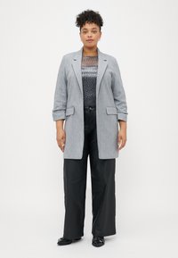 Grijze blazer van gestructureerde stof met revers, gecombineerd met een met patroon shirt en brede zwarte broek, afgemaakt met zwarte schoenen.