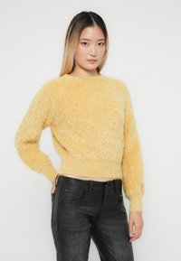 Jonge vrouw in een donzige gele trui en zwarte, gestructureerde broek, staand met één hand in de zak tegen een effen witte achtergrond.