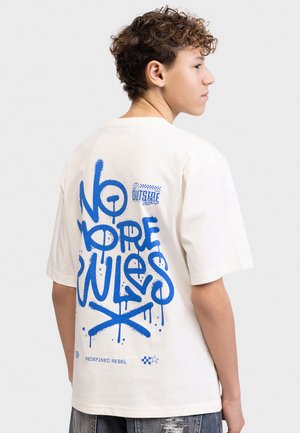 Έφηβος με σγουρά μαλλιά που φοράει λευκό T-shirt με μπλε γκράφιτι κείμενο "No More Rules" και "Redefined Rebel" στην πλάτη, στραμμένος στο πλάι.