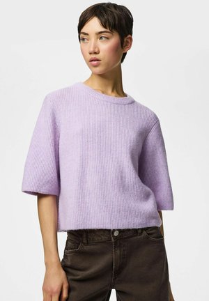Femme aux cheveux courts et foncés, portant un pull en maille lilas à manches courtes et un pantalon foncé, regardant vers sa gauche sur un fond uni.