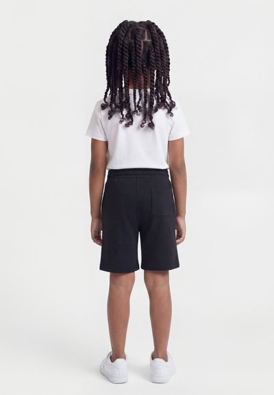 Παιδί με μεσαίο καφέ δέρμα και μακριές πλεξούδες στραμμένες προς τα πίσω, φοράει λευκό μπλουζάκι, μαύρο σορτς και λευκά αθλητικά παπούτσια.