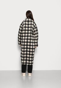 Dlouhý, přehnaně velký černobílý kostkovaný kabát s jemnou teksturou. Obsahuje spadlé ramena a široký límec, v kombinaci s černými kalhotami a béžovými botami.
