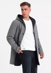 Abrigo gris de un material similar a la lana con capucha negra, con botones en la parte delantera, dos bolsillos laterales y un corte relajado combinado con una camisa blanca y jeans oscuros.