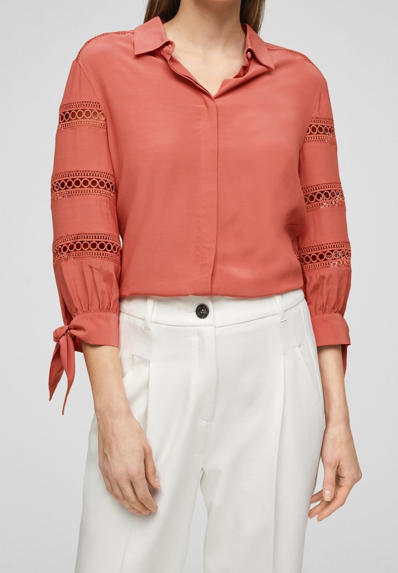 Kobieta ubrana w bluzkę w kolorze rdzy z koronkowymi wycięciami na rękawach i wiązanymi mankietami, w połączeniu z białymi spodniami z wysokim stanem i zapięciem na guziki.