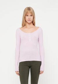 Top de manga longa em rosa claro com textura canelada, com um decote redondo e cinco botões prateados ao centro, combinado com calças ajustadas em verde oliva.