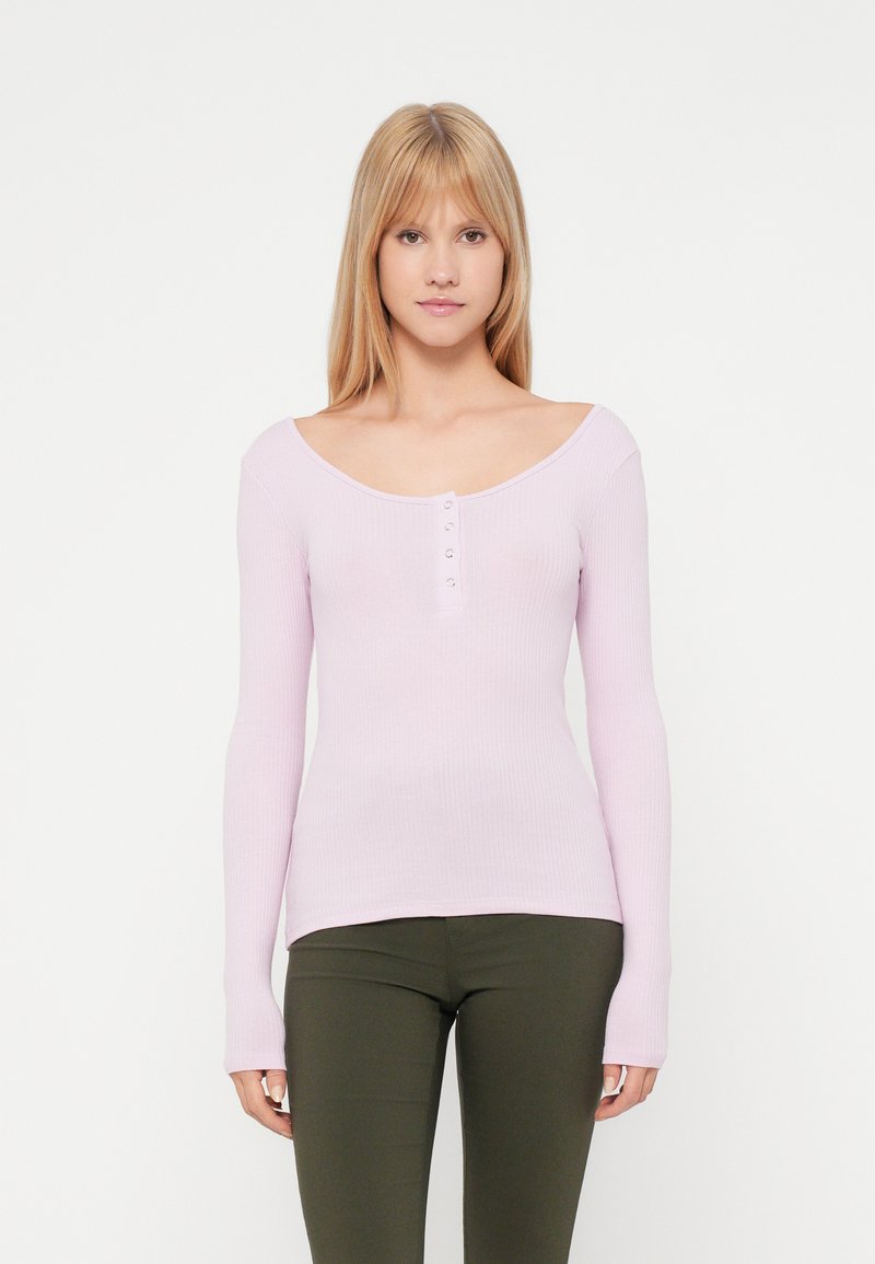 Top de manga longa em rosa claro com textura canelada, com um decote redondo e cinco botões prateados ao centro, combinado com calças ajustadas em verde oliva.