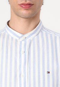 Camicia a righe blu chiaro e bianca con colletto alto, dotata di due bottoni bianchi e un piccolo logo ricamato sul petto.
