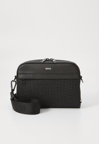 Sac à bandoulière noir en matériau texturé, doté d'une fermeture éclair, d'un logo à l'avant et d'une bandoulière amovible. Forme rectangulaire compacte.