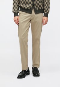 Pantaloni chino beige dal taglio dritto, realizzati in tessuto liscio. Abbinati a mocassini neri in pelle e a un cardigan scuro con motivo.