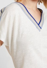Lehký, krémový top s hlubokým výstřihem do V, který má modré a metalické akcenty kolem výstřihu a volné krátké rukávy.