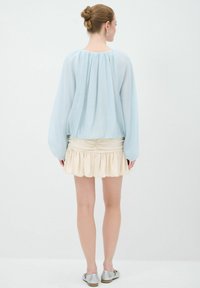 Himmelblaues, locker sitzendes Blusenshirt mit gerafftem Ausschnitt; kombiniert mit einem cremefarbenen, rüschengesäumten Minirock; das Model trägt silberne Ballerinas; minimalistisches Design.