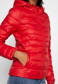 Veste matelassée rouge avec capuche, dotée d'un design matelassé, fermeture éclair à l'avant et poignets élastiqués. Comprend des poches latérales et une texture de tissu brillante.