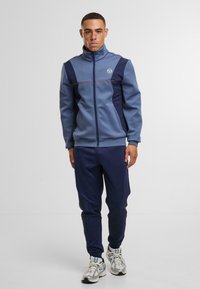 Tepláková souprava v modré a námořnické barvě, vybavená vysokým límcem, zipovým zapínáním a kontrastními panely. Doplněná světle šedými teniskami.