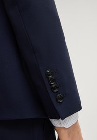 Blazer blu navy in tessuto strutturato; presenta quattro bottoni neri sul polsino e una camicia bianca con motivo geometrico blu sotto.