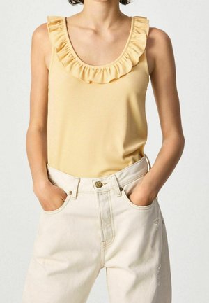 Mujer con una blusa amarilla sin mangas con volantes metida en pantalones de talle alto de color blanco roto, con las manos en los bolsillos.