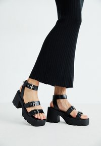 Sandalias de plataforma negras con suelas gruesas y dentadas, y múltiples correas, que presentan hebillas de metal y una textura similar al cuero suave.
