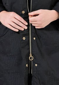Černá bunda s texturou, zlatými doplňky a zipovým uzávěrem. Obsahuje dvě boční kapsy a lesklý povrch.