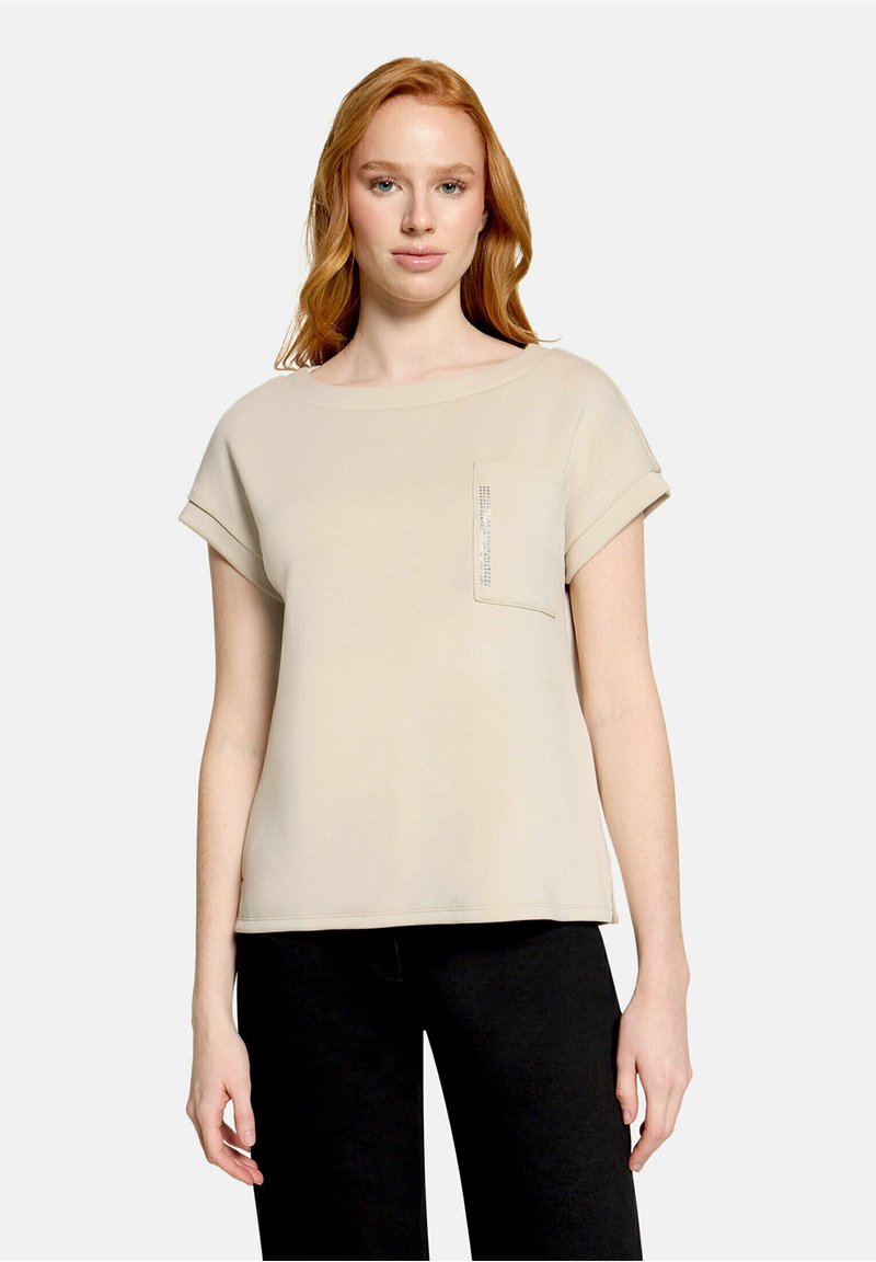 Jeune femme aux cheveux roux clair portant un haut beige à manches courtes avec une poche avant et un pantalon noir sur fond uni.